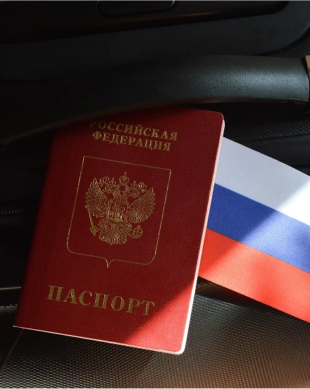 A Russian passport lies open on a black suitcase, partially obscured by a Russian flag.